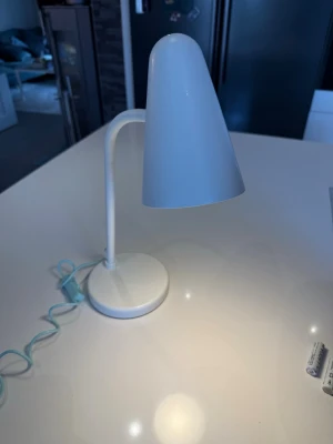 Vit och ljusblå skrivbordslampa - Snygg skrivbordslampa i vit metall med konformad ljusblå lampskärm. Lampan har rund fot och böjd arm, samt en matchande ljusblå sladd med strömbrytare. Perfekt för plugghörnan eller som läslampa.