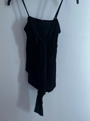 Svart topp med knyt framtill - Trendig svart topp med smala axelband och knyt framtill som ger en snygg detalj. Toppen har asymmetrisk nederkant och är gjord i ett mjukt, tunt tyg som känns skönt mot huden. Perfekt för dig som gillar en stilren men ändå unik look. Aldrig använd