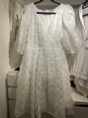 Vit omlottklänning med rosdetaljer - Romantisk vit omlottklänning från Bubbelroom med v-ringning och puffiga trekvartsärmar. Klänningen har ett vackert rosformat mönster i tyget och en lätt utsvängd kjol. Perfekt för sommarens speciella tillfällen och speciellt för dig som tar studenten! 