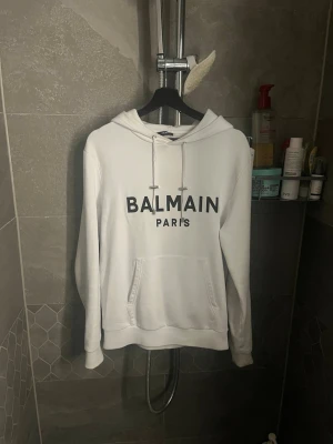 Vit hoodie från Balmain Paris - Snygg vit hoodie från Balmain Paris med stor svart logga framtill. Klassisk modell med huva och dragsnören samt en stor magficka. Tillverkad i mjukt material som känns skönt mot huden. Perfekt för en avslappnad och trendig look.