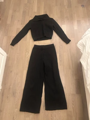 Svart ribbat loungewear-set - Mysigt svart loungewear-set med långärmad, croppad tröja med krage och vida ribbade byxor med resår i midjan. Perfekt för chill dagar hemma eller när du vill vara bekväm men ändå se snygg ut. Materialet är stickat och mjukt.
