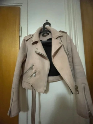 Ljusrosa skinnjacka med bälte - Snygg ljusrosa skinnjacka i bikerstil med silvriga dragkedjor, axelklaffar och bälte i midjan. Jackan har en croppad passform och coola detaljer som ger en edgy look. Perfekt för dig som vill sticka ut med en unik färg på klassisk modell.