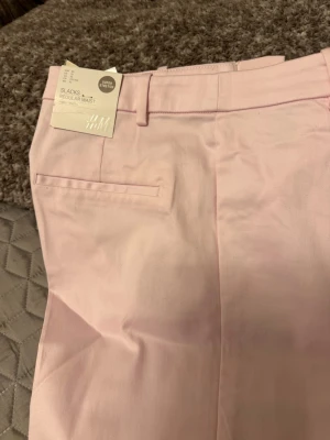 Ljusa rosa kostymbyxor H&M - Snygga ljusrosa kostymbyxor från H&M med regular waist och raka ben. Byxorna har bälteshällor, diskreta bakfickor och är gjorda i ett mjukt, vävt material. Perfekta för en clean och stilren look.