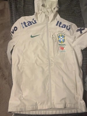 Vit Nike Brasil jacka med huva - Säljer en vit sportjacka från Nike med huva och dragkedja. Jackan har Brasil landslagslogga, grönt Nike-märke och flera sponsorloggor i blått på bröst och ärmar. Perfekt för dig som gillar fotboll och vill ha en snygg och funktionell jacka.
