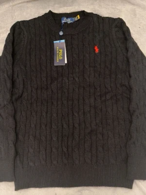 Svart kabelstickad tröja Polo Ralph Lauren - Kabelstickad svart tröja från Polo Ralph Lauren med klassisk röd logga broderad på bröstet. Tröjan har rund halsringning och ribbade muddar vid ärmslut och nederkant. Perfekt för dig som gillar stilrena och tidlösa plagg.