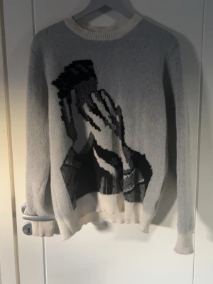 Grå stickad tröja med grafiskt motiv - Stickad tröja i ljusgrått med ett stort svartvitt grafiskt motiv framtill som föreställer händer. Tröjan har rund halsringning och ribbade muddar vid ärmslut och nederkant. Perfekt för dig som gillar unika plagg med konstnärlig touch.