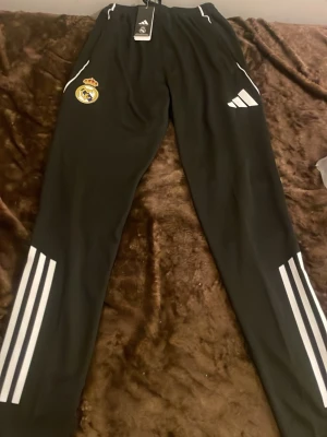 Svarta Real Madrid träningsbyxor Adidas - Säljer ett par svarta träningsbyxor från Adidas med Real Madrid-logga på ena benet och klassiska vita ränder längs benen. Byxorna har elastisk midja och är perfekta för fotbollsträning eller chill. Adidas-logga på andra benet.