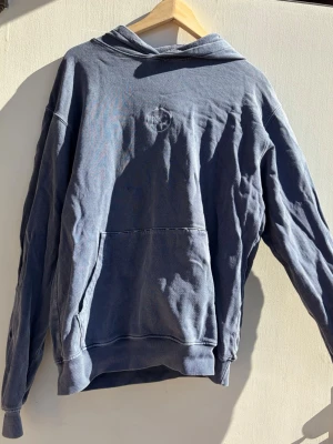 Blå hoodie från Noon Goons - Säljer en blå hoodie från NG med diskret logga på bröstet. Hoodien har en klassisk känguruficka framtill och huva. Tillverkad i mjukt bomullsmaterial och har en avslappnad passform, perfekt för en chill look.