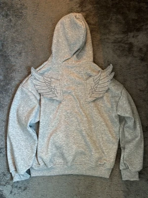 Grå hoodie med vingar  - Jätte snygg grå hoodie med vingar. Ena vingen blir lite ”floppig” eftersom som har ingenting att stanna uppe med när man har den på sig. Storlek M. Liknar jeremy scott x adidas hoodien och är också jätte mjuk på insidan. Har använts en gång där jag provade den. Har inget märke på sig. Pris kan diskuteras💗💗