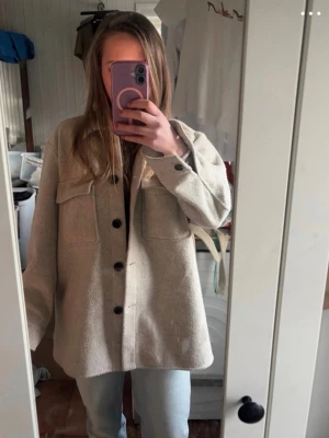 Beige overshirt från Dobber - Snygg beige overshirt från Dobber med stora bröstfickor och svarta knappar framtill. Overshirten har en rak passform och är gjord i ett mjukt, ull-liknande material. Perfekt att slänga över en hoodie eller t-shirt för en chill look.