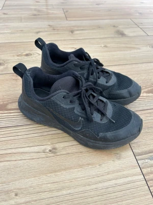 Svarta Nike sneakers - Helsvarta Nike sneakers med låg profil. Ovandelen är i mesh och syntetmaterial för bra andningsförmåga och komfort. Klassisk snörning och diskret Nike-logga på sidan. 