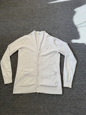 Beige cardigan från J.Lindeberg - Passar M. Stilren beige cardigan från J.Lindeberg med v-ringning och knäppning framtill. Tröjan har två fickor och diskret logga på bröstet. Tillverkad i tunt, mjukt material som passar perfekt för lager på lager och en clean look.