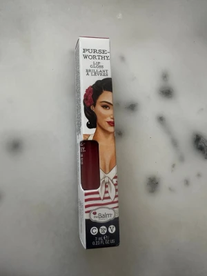 theBalm Purse-Worthy Lip Gloss Wristlet - Snyggt läppglans från theBalm i nyansen Wristlet. Färgen är en djup röd med glansig finish. Kommer i en stilren förpackning med retrodesign och innehåller 7 ml. Perfekt för dig som vill ha ett glossigt resultat med färg som sticker ut.