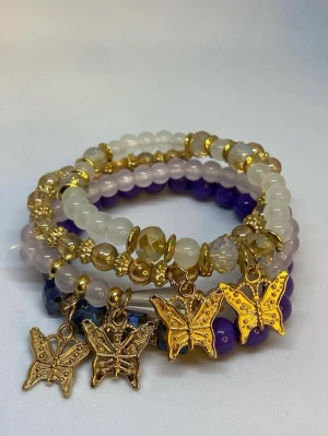 Armband med pärlor och fjärilar - Flera elastiska armband med pärlor i vitt, lila och guldtoner. Varje armband har guldfärgade detaljer och fjärilsberlocker som hänger ner. Pärlorna är runda och facetterade, vilket ger ett glittrande intryck. Ej använd utan kommer från min small business.