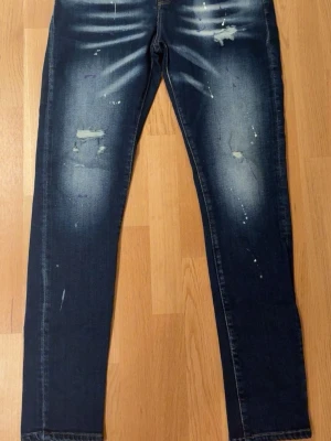 Mörkblå slitna skinny jeans - Säljer ett par mörkblå skinny jeans med slitningar och blekta detaljer framtill. Jeansen har klassisk femficksmodell, knappgylf och coola färgstänk för en trendig look. Perfekta för dig som gillar streetstyle och vill sticka ut lite extra.