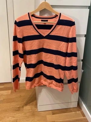 Randig stickad tröja Tommy Hilfiger - Snygg stickad tröja från Tommy Hilfiger i persikoorange med breda mörkblå ränder. Tröjan har V-ringning och långa ärmar, perfekt för en avslappnad men ändå stilren look. Materialet känns mjukt och lätt, vilket gör den skön att bära.
