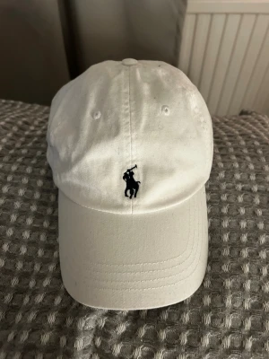 Vit keps från Polo Ralph Lauren - Säljer en stilren vit keps från Polo Ralph Lauren med klassisk böjd skärm. Framtill finns den ikoniska broderade polospelaren i marinblått och baktill står det 'POLO' broderat. Kepsen är i bomull och har justerbar rem bak.