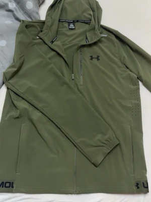 Olivgrön jacka  från Under Armour - Snygg olivgrön jacka  från Under Armour med dragkedja framtill och huva. Jackan  har långa armar, diskret logga på bröstet och ventilerande hål på sidorna. Perfekt för träning eller chill. Materialet känns lätt och stretchigt. En byxa som tillhör den också följer med 