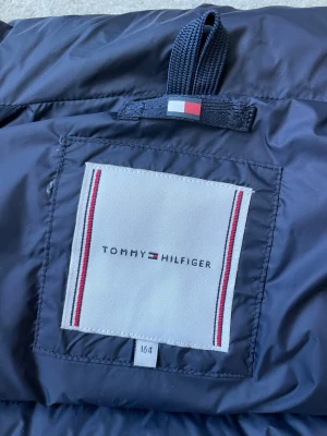 Mörkblå Tommy Hilfiger lättviktsjacka i mycket bra skick  - Mörkblå lättviktsjacka från Tommy Hilfiger med quiltad design och hög krage. Jackan har tydliga logodetaljer på både insidan och baksidan av kragen. Materialet är glansigt och jackan är perfekt för vår och höst.