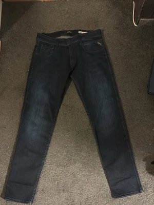 Mörkblå slim fit jeans från replay - Snygga mörkblå slim fit jeans från replay med klassisk femficksdesign och diskreta slitningar framtill. Jeansen har raka ben och är tillverkade i ett mjukt denimtyg som ger en bekväm passform. Perfekta för en stilren och avslappnad look.