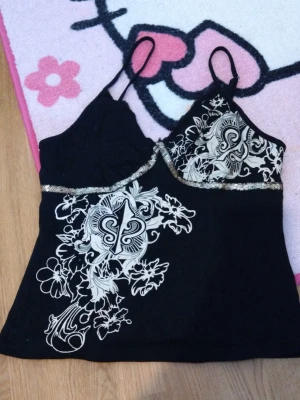 Svart linnetopp med floral tryck - Snyggt svart linne med tunna och justerbara axelband och dekorativt blommigt/floralt mönster i pastel gul framtill. Toppen har glittriga paljetter under bysten som ger extra stil. Perfekt för dig som vill ha något unikt och trendigt i garderoben. Ett litet hål som går att fixa enkelt med tråd och nål. 🥰😊🤫❤️ Vi kan diskutera priset 💸 💵 