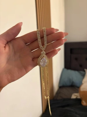 Guldigt halsband med droppformad kristall - Snyggt halsband i guldton med dubbla kedjor och en stor droppformad kristall i mitten, omgiven av glittrande stenar. Hängande kedjor under hänget ger extra bling. Perfekt för dig som vill sticka ut med accessoarer som verkligen syns.
