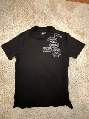 Svart t-shirt från Mercier  - Snygg svart t-shirt från Mercier med flera broderade patches och logotyper på bröstet. Klassisk rund hals och korta ärmar. Tillverkad i mjuk bomull som känns skön mot huden. Perfekt för dig som gillar streetwear och vill sticka ut lite extra.