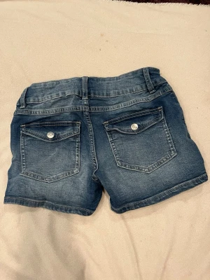 Blå lågmidjade jeansshorts med fickor - Säljer ett par klassiska blå jeansshorts från denim day. dubbla knappar fram och snygga bakfickor med. Perfekta för varma sommardagar.