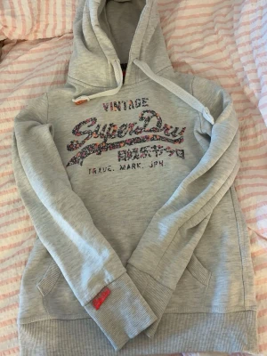 Grå Superdry hoodie med blommigt tryck - Snygg ljusgrå hoodie från Superdry med blommigt Superdry-tryck på bröstet och japanska tecken. Klassisk huva med vita snören och känguruficka framtill. Mjuk bomullsblandning och ribbade muddar vid ärmar och nederkant. Perfekt för en avslappnad stil. Super snygg till ett par jeans och snygga skor!💓