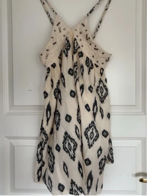 Mönstrad kortklänning - Säljer en ljus kortklänning med svart mönster och tunna axelband. Klänningen har dekorativa pärlor upptill och ett boho-inspirerat mönster. Materialet känns som bomull och den har en avslappnad passform, perfekt för sommardagar.