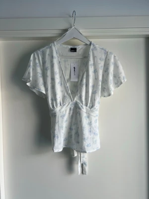 Vit blommig omlottopp från Gina tricot  - Söt helt nya vit omlottopp från Gina tricot med ljusblått blommönster. Toppen har korta ärmar, v-ringning och knytband i midjan för en feminin siluett. Perfekt för vår och sommar, till jeans eller kjol.