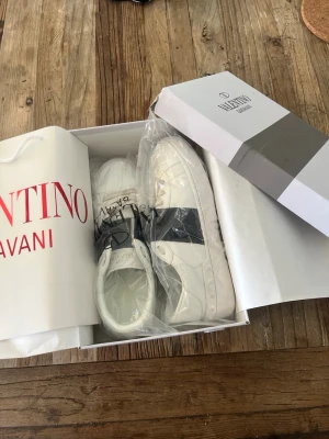 Valentino vita sneakers med svart detalj - Snygga vita sneakers från Valentino Garavani med en bred svart rem över sidan och diskret logga. Skorna har klassisk låg profil, rund tå och är tillverkade i slätt läder. Perfekta för dig som vill ha en clean och exklusiv look. Använda typ 2 gånger men de är lite för små för mig hör av er vid frågor kan sänka priset vid snabb affär ✌🏻