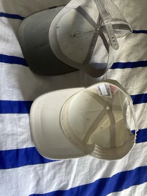 Två kepsar: Adidas & Pug-motiv - Säljer två kepsar. En beige Adidas-keps med vit logga framtill och justerbar rem bak. Den andra är en brun och beige truckerkeps med mesh och ett broderat pug-motiv på framsidan. Båda har böjd skärm och klassisk passform.