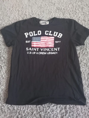 Svart t-shirt från Saint Vincent - Säljer en svart t-shirt från Saint Vincent med tryck av amerikanska flaggan och texten 'POLO CLUB SAINT VINCENT U.S OF A CREW LEGACY' på bröstet. Klassisk passform och rund hals. Perfekt för dig som gillar sportig och stilren look.