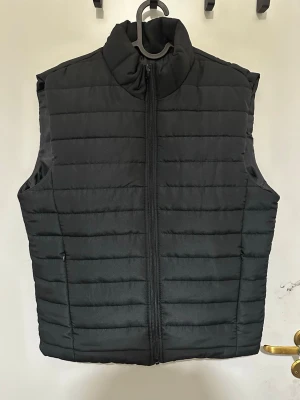 Herr väst  - Stilren svart dunväst med quiltat mönster och dragkedja framtill. Västen har hög krage och två sidofickor med dragkedja. Perfekt att slänga över en hoodie eller tröja för extra värme. Materialet är lätt och smidigt, vilket gör den enkel att bära.