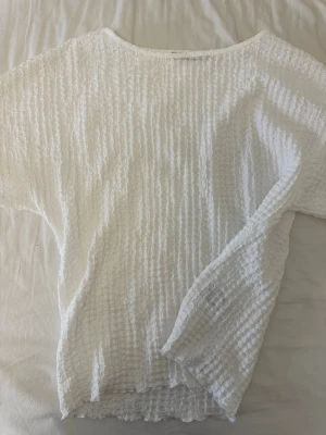 Vit strukturerad topp med kort ärm - Säljer en vit topp med kort ärm och vågig, strukturerad yta. Toppen har en lös passform och är tillverkad i ett lätt, luftigt material som passar perfekt till varma dagar. Enkel och stilren design som är lätt att matcha.