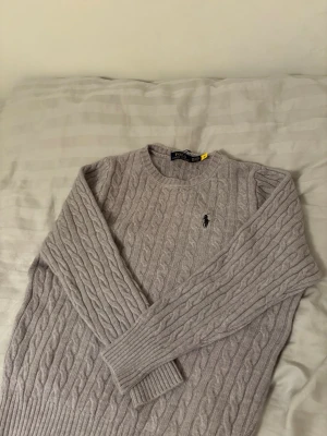Stickad tröja Polo Ralph Lauren - Säljer en ljusgrå kabelstickad tröja från Polo Ralph Lauren med rund halsringning, lite liten i storlek skulle jag säga.