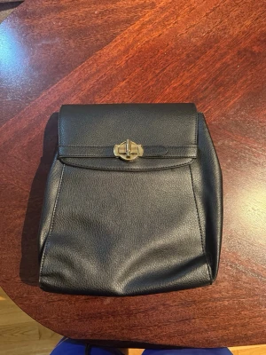Svart handväska i skinnimitation - Stilren svart handväska i skinnimitation med lock och snyggt guldfärgat vridlås. Väskan har en enkel, rektangulär form och passar perfekt till dig som gillar minimalistisk design. Praktisk storlek för det viktigaste och lätt att matcha med olika outfits. Köpt i JUSTFAB. Helt ny utan prislapp.