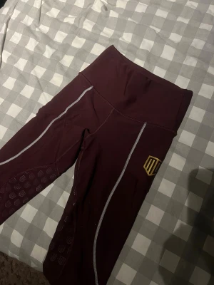 Vinröda ridbyxor från maya delorez - Snygga vinröda ridbyxor från maya delorez med hög midja och vita kontrastsömmar. De har ett gult logotryck på benet och mönstrade detaljer på underbenen. Materialet är stretchigt och passar perfekt till träning eller chill.