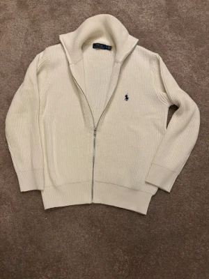 Vit ziptröja från Polo Ralph Lauren - Klassisk vit ribbstickad ziptröja från Polo Ralph Lauren med hög krage och blå broderad logga på bröstet. Tröjan har långa ärmar, dragkedja framtill och breda muddar vid ärmslut och nederkant. Perfekt för en clean och stilren look.