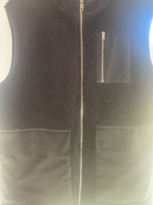 Svart dunväst från Ciszere - Stilren svart dunväst från Ciszere med mjuk teddystruktur, hel dragkedja framtill och tre fickor – två stora nedtill och en bröstficka med dragkedja. Västen har detaljer i tyg och passar perfekt för lager-på-lager-look.