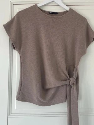 Beige topp med knyt från Zara - Säljer en beige topp från Zara med korta ärmar och snygg knytning i sidan. Toppen har en mjuk, stickad känsla och rund halsringning. Perfekt att styla med jeans eller kjol för en clean look.