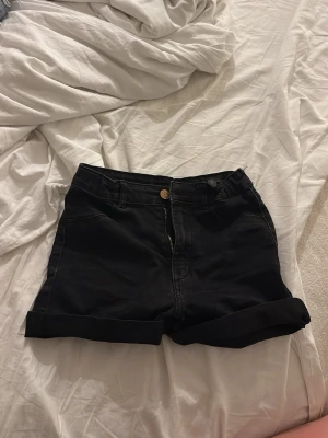 Svarta low waist jeansshorts - Säljer ett par svarta low waist jeansshorts med uppvikta benslut. Shortsen har klassisk femficksdesign, knapp och dragkedja framtill samt bälteshällor. Perfekta till sommaren och lätta att matcha med olika stilar.