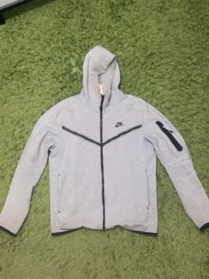 Grå Nike hoodie med dragkedja - Snygg grå hoodie från Nike med hel dragkedja framtill och svart Nike-logga på bröstet. Hoodien har svarta detaljer, fickor med dragkedja och en extra ficka på ärmen. Tillverkad i mjukt material och har en huva med dragsko.