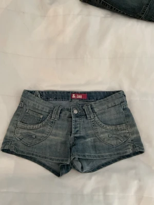 Blå jeansshorts från Chio - Säljer ett par blå lågmidjade jeansshorts från Chio med låg midja och klassisk femficksdesign. Shortsen har slitna detaljer, bälteshällor och knappgylf. Bakfickorna har broderade detaljer och passformen är tight och kort.