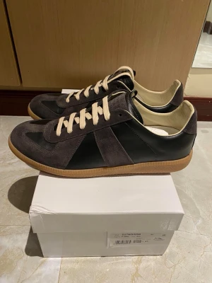 Maison Margiela Gats  - Snygga sneakers från Maison Margiela med grå mockadetaljer och svarta partier i skinn. Skorna har beige snörning och klassisk gummisula. Perfekt för dig som gillar stilrena och tidlösa sneakers med en modern twist.