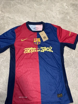Barça x Nike matchtröja blå/röd - Snygg FC Barcelona matchtröja från Nike i klassiska blå och röda färger. Tröjan har klubbmärke på bröstet, gul Nike-logga och trycket 'Cactus Jack' i gult. Tillverkad i lätt, ventilerande Dri-FIT-material som passar perfekt till fotboll eller träning.