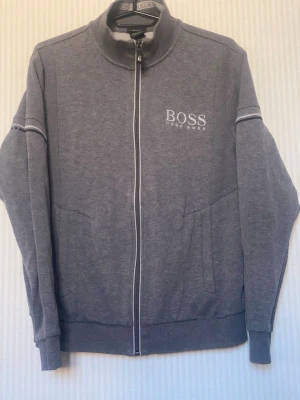 Grå ziptröja från BOSS Hugo Boss - Snygg grå ziptröja från BOSS Hugo Boss med vit logga på bröstet och vita detaljer längs dragkedjan. Tröjan har hög krage, långa ärmar och ribbade muddar. Perfekt till chill eller streetwear-stil.