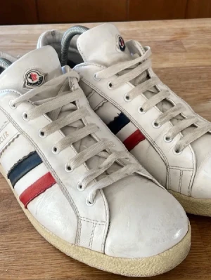 Vita Moncler sneakers med ränder - Snygga vita sneakers från Moncler i läder med klassiska röda, vita och blå ränder på sidan. Skorna har lågt skaft, vita snören och gummisula. Moncler-logga på plösen och sidan. Perfekta för dig som gillar stilrena och sportiga skor.