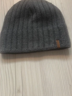 Mörkgrå ribbstickad mössa - Säljer en stilren mörkgrå ribbstickad mössa med diskret logga på en brun läderlapp. Mössan har en klassisk form och är tillverkad i mjukt material som håller dig varm under kalla dagar.